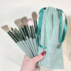 A hand holds a set of thirteen professional makeup brushes with green handles and a teal fabric brush bag.