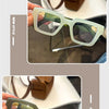 Paire de lunettes à monture carrée rétro vert clair avec une sangle en cuir marron, posées sur une surface blanche.