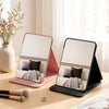 A foldable portable makeup mirror in black and pink rests on a wooden table next to dried flowers.