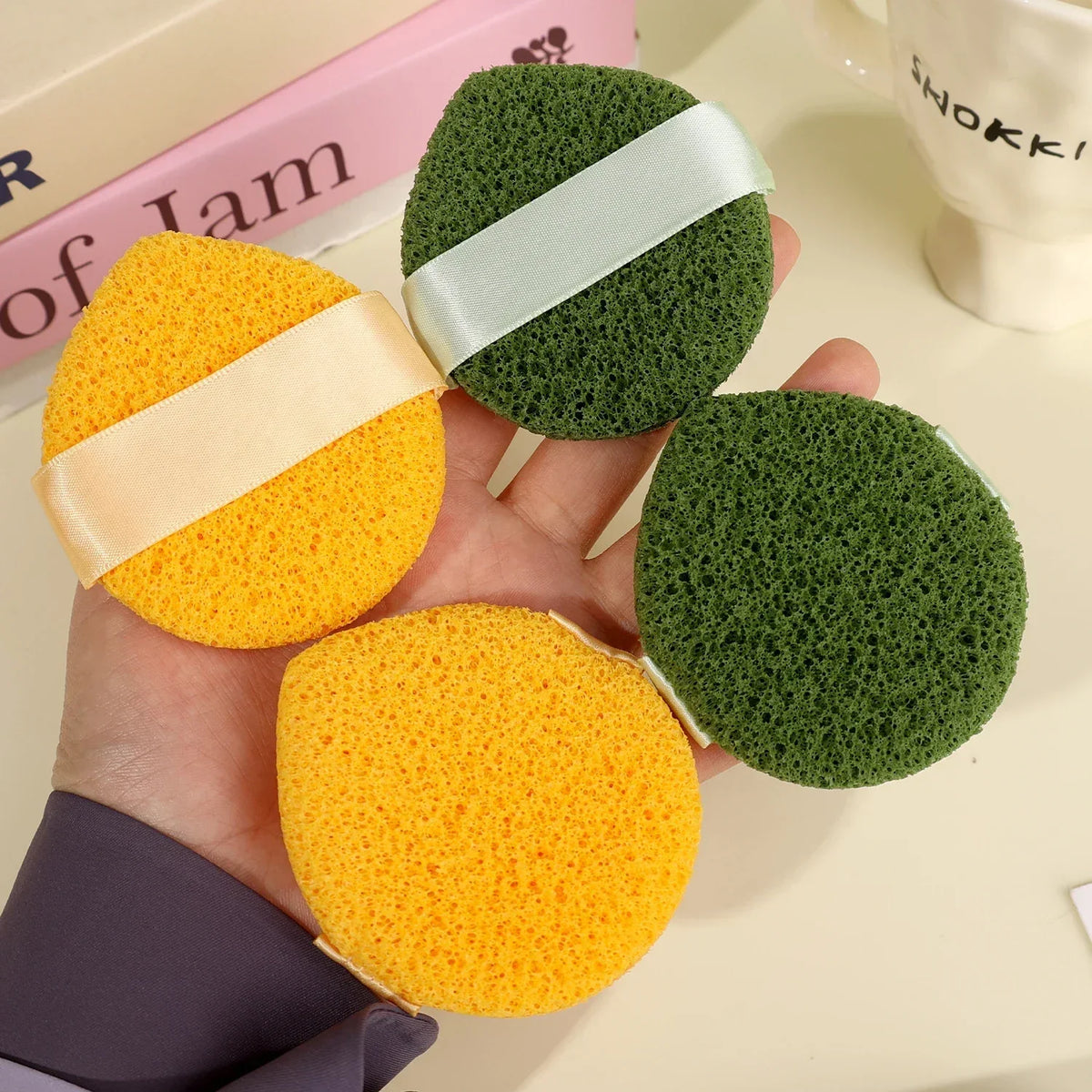 Two exfoliating cleansing sponges, one yellow and one green, are held in a hand.