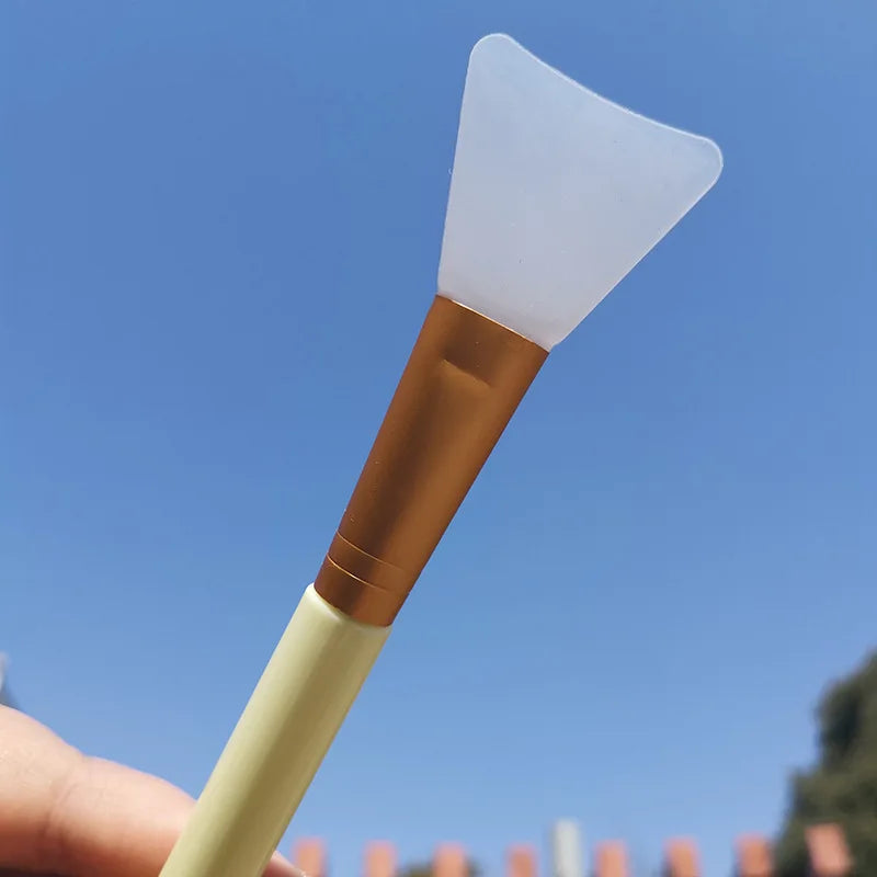 A silicone facial mask applicator brush with a white silicone tip and a brown handle is held against a blue sky background.