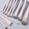 A collection of thirteen professional makeup brushes in a pale pink and taupe color palette arranged diagonally on a light gr