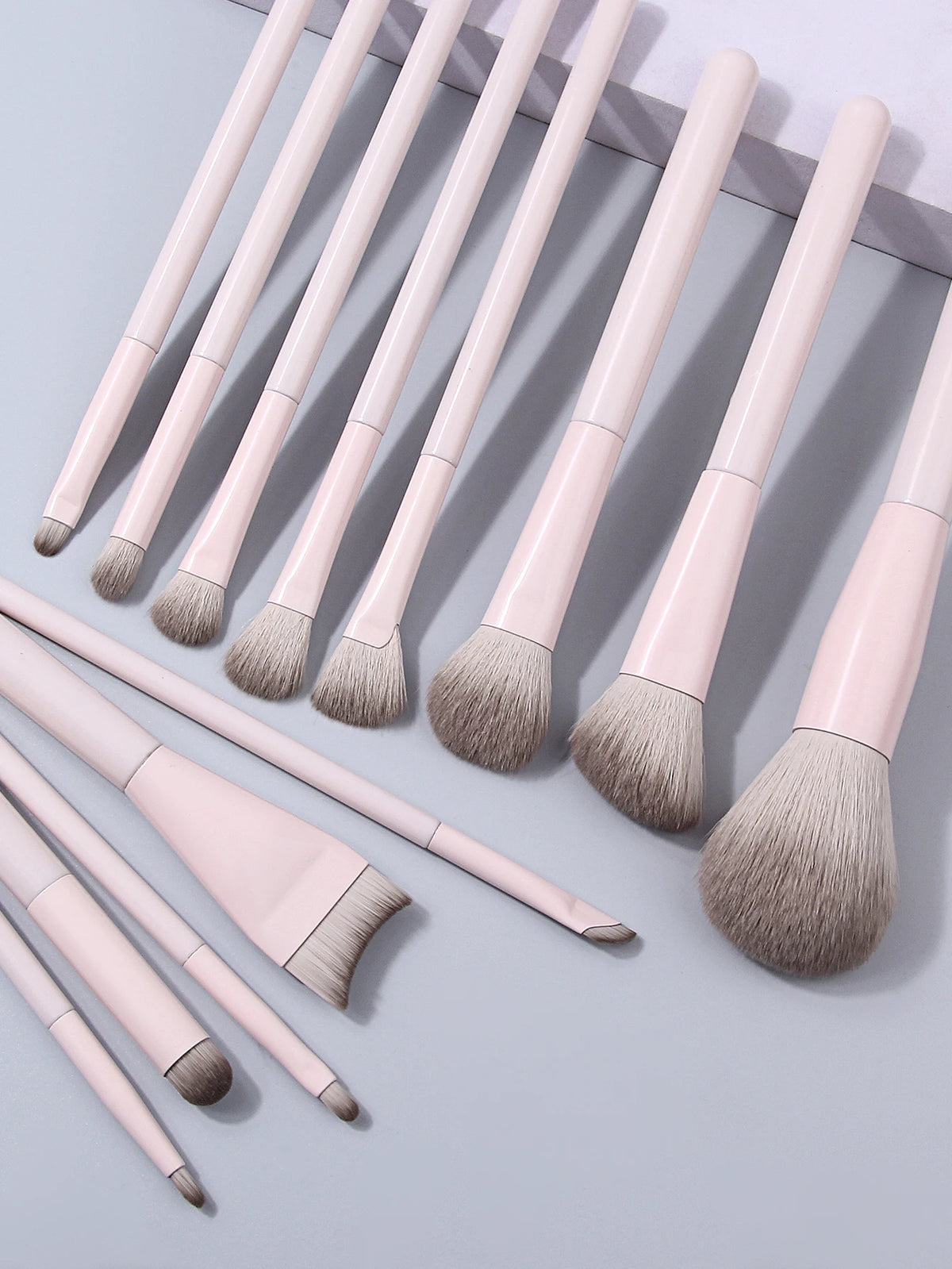 A collection of thirteen professional makeup brushes in a pale pink and taupe color palette arranged diagonally on a light gr