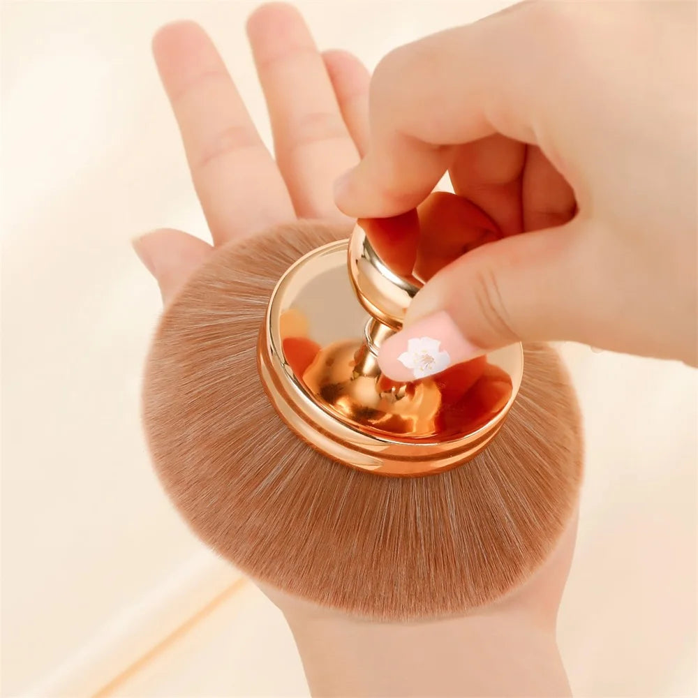 Une main tient un pinceau de maquillage kabuki extra-large avec un manche doré et des poils bruns souples.