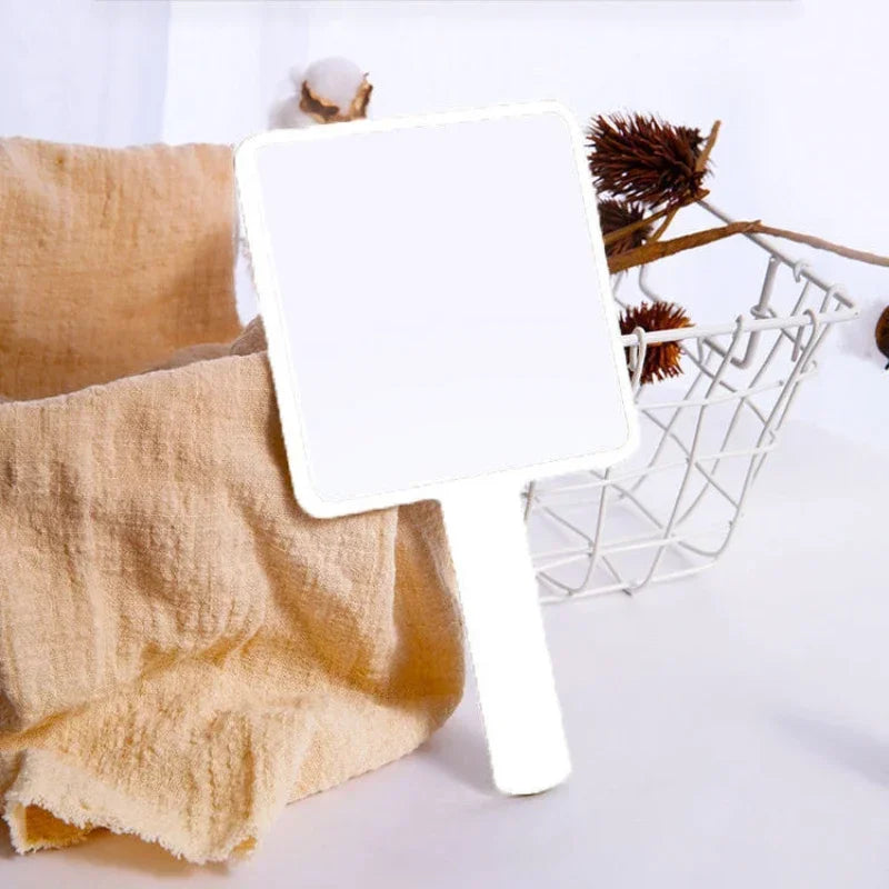 Miroir de maquillage portable blanc avec poignée, à côté d'une serviette beige sur une surface blanche.