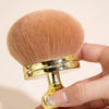 A golden makeup brush with soft, light brown bristles is held in a hand.