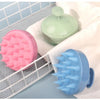 A pink silicone scalp massage brush and a blue silicone body brush are displayed on a white towel within a wire rack.