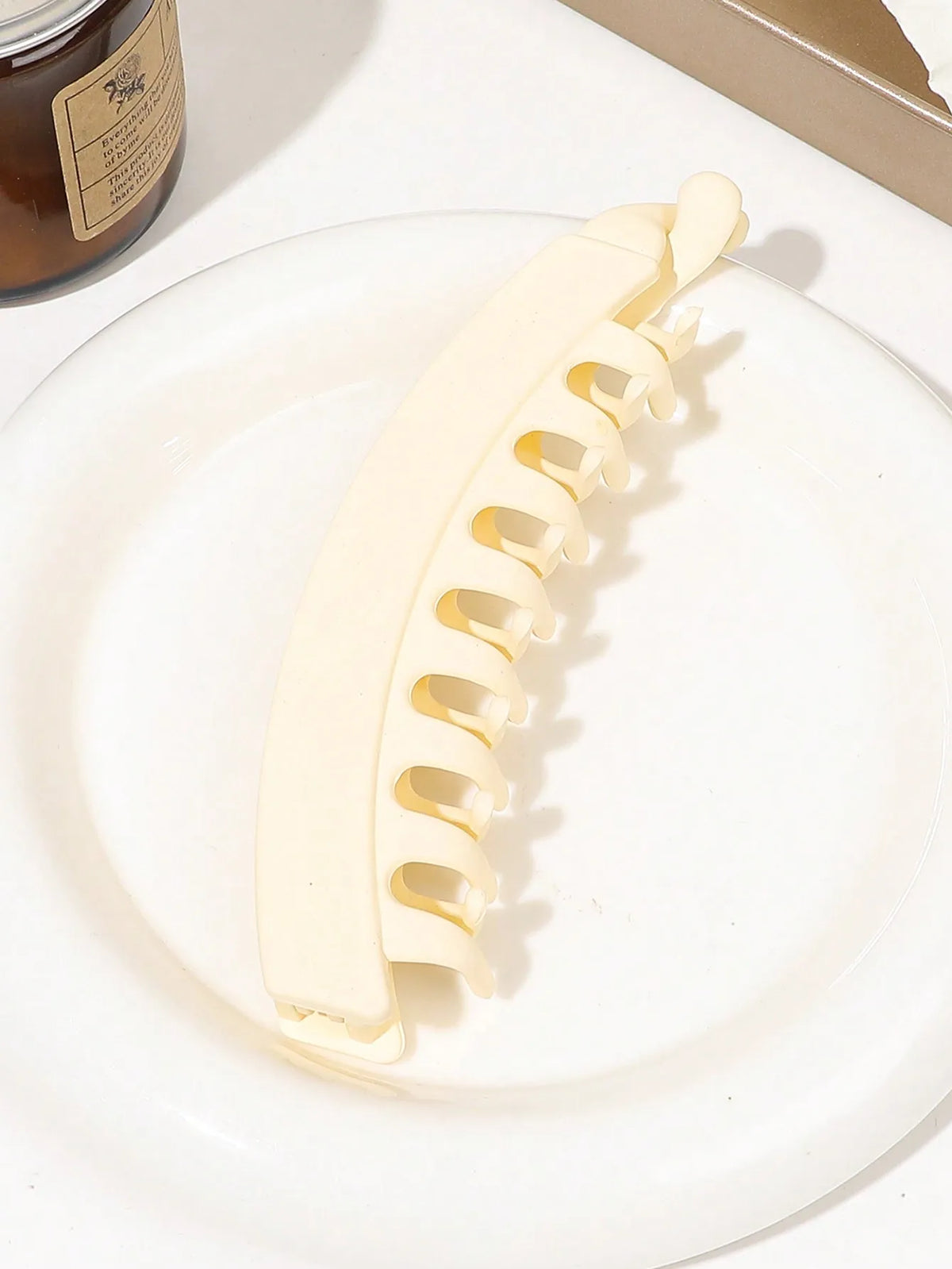 A white banana hair clip is shown on a white plate, next to a jar.