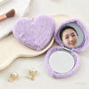 A purple heart-shaped foldable travel makeup mirror with a reflective surface and pearl-like jewelry is displayed on a light 