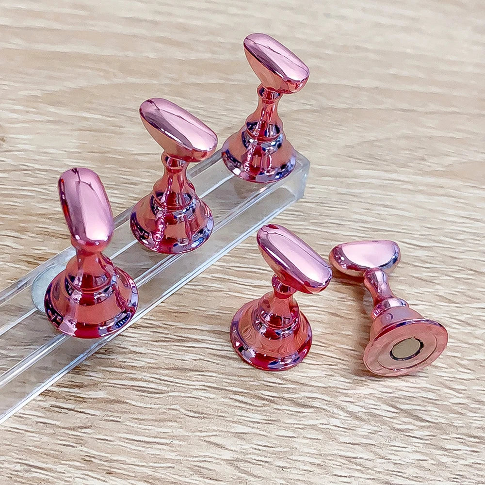 Un ensemble de supports à ongles magnétiques roses métalliques avec des boutons décoratifs, présentés dans un support en acry