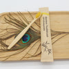 A bamboo toothbrush packaged in paper is displayed on a wooden board with a background of feathers.