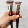 Une main tient deux pinceaux de maquillage pour fond de teint liquides inclinés avec des poils roses et violets.