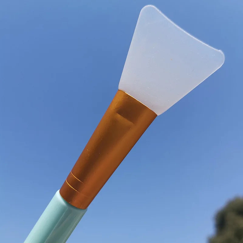 A silicone facial mask applicator brush with a white silicone pad and a copper-colored handle against a blue sky.