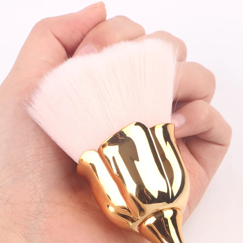 A hand holds a gold-colored nail art dust brush set with a pink feather brush.