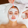 A woman is applying a white facial mask to her face using a pink silicone applicator under a white towel.