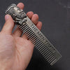 A hand holds a silver beard and mustache comb with a sculpted face design.