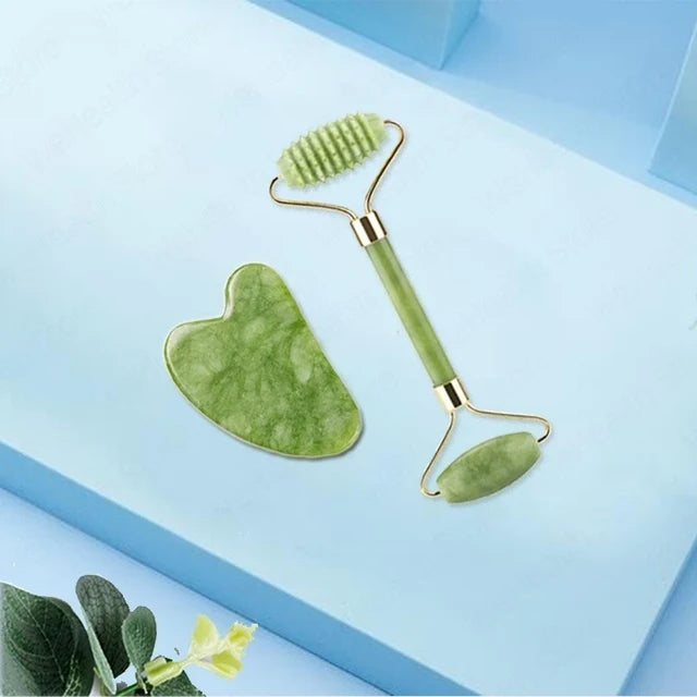 A jade gua sha facial massager tool with a green heart-shaped stone and a metal handle is shown on a light blue surface.