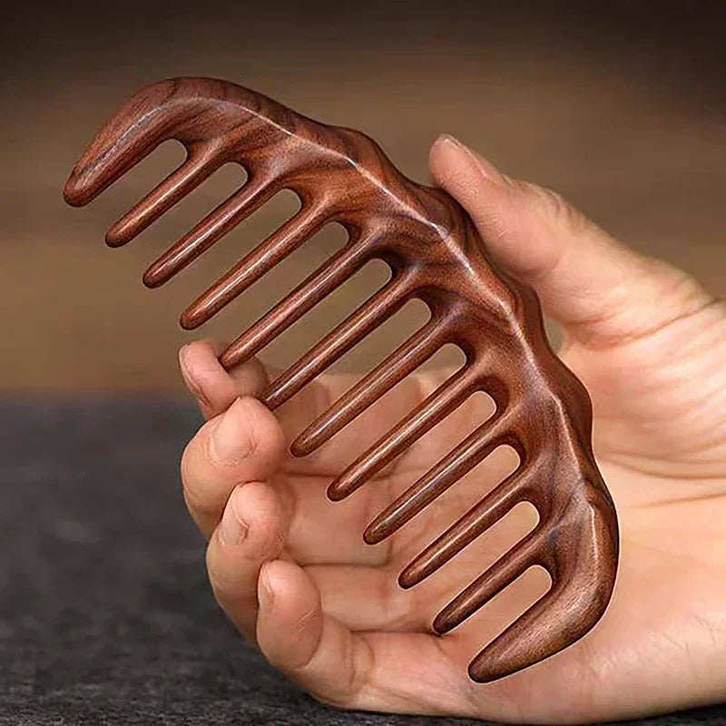 A hand holds a natural golden sandalwood wide-tooth detangling comb with curved teeth.