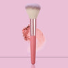 A pink face powder brush with a fluffy white synthetic hair and loose pink powder surrounding the handle.