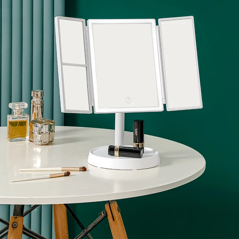 A white foldable LED makeup mirror with three panels is displayed on a round table against a dark green wall.
