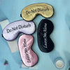 Three silk sleep masks are displayed on a light blue surface, featuring embroidered text like Do Not Disturb and Leave Me Alo