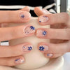 A hand displays blue floral press-on false nails on fingers against a light background.