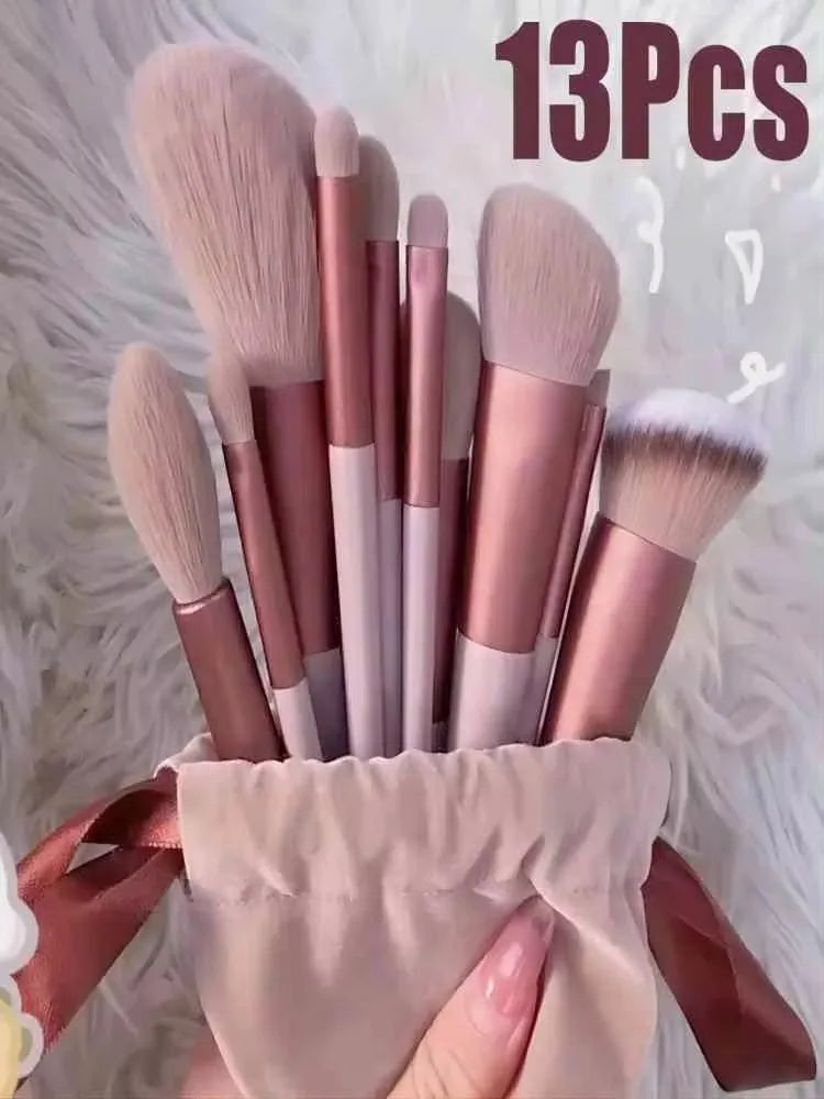 A collection of 13 professional makeup brushes in various shades of pink and nude are displayed on a white fluffy surface.