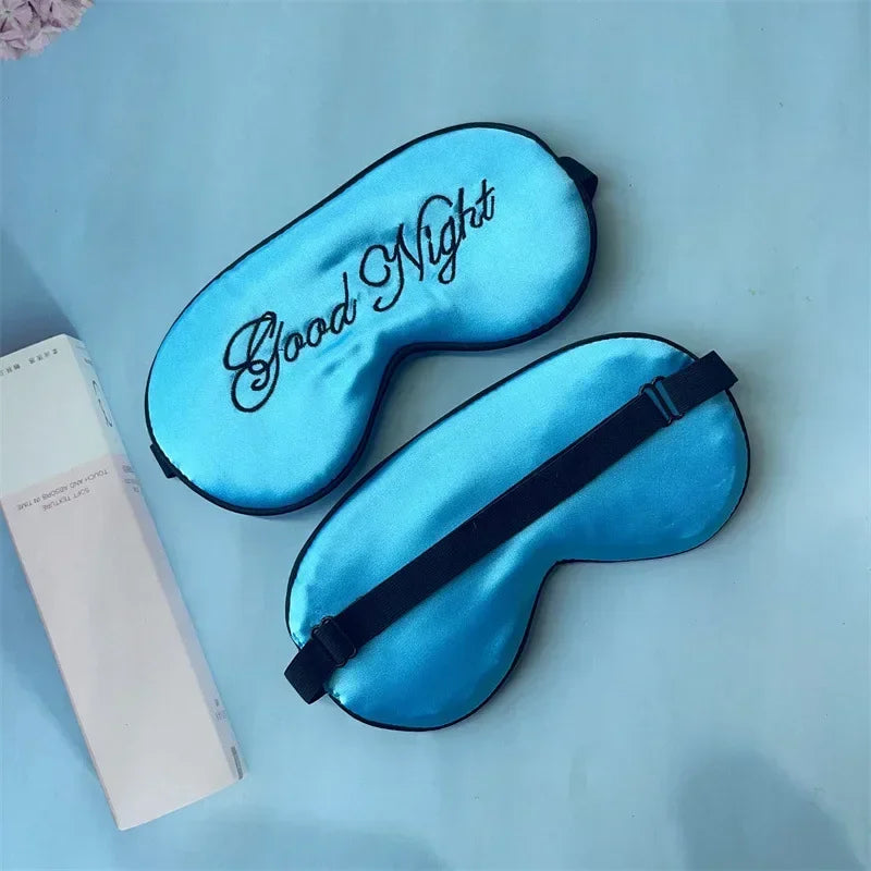 A bright blue silk sleep mask embroidered with the text Good Night is displayed next to its packaging.