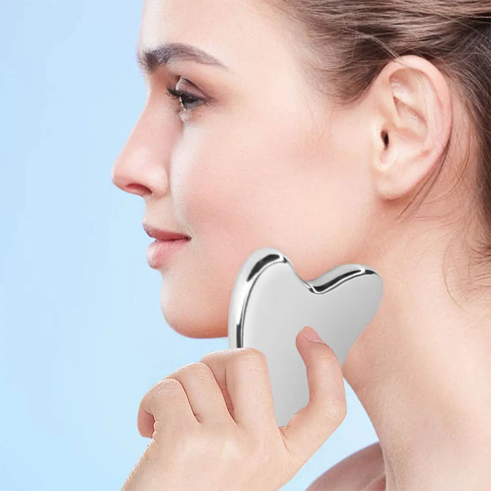 A woman is using a silver stainless steel gua sha tool on her cheekbone for facial massage.