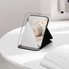 A black foldable portable makeup mirror with a white reflective surface is shown on a white table.