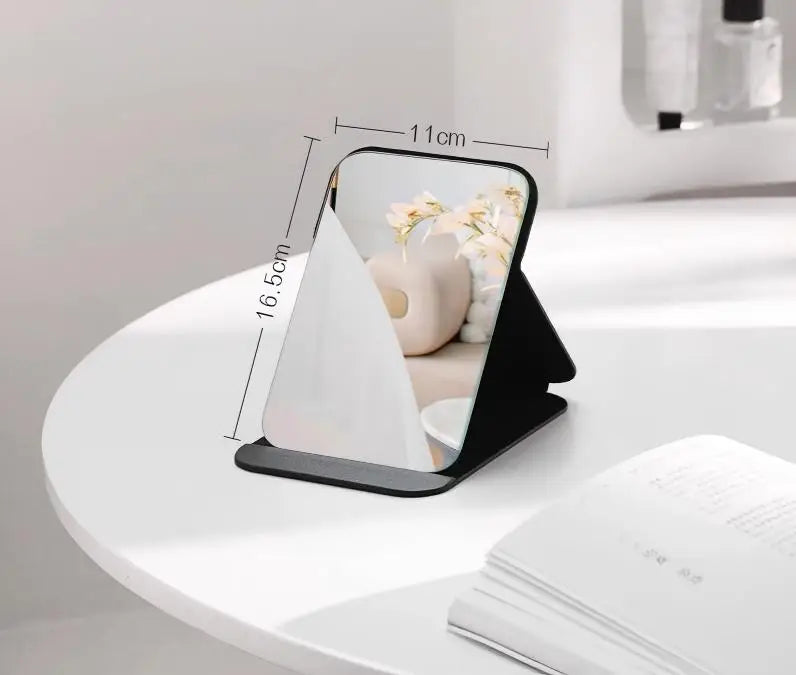 A black foldable portable makeup mirror with a white reflective surface is shown on a white table.