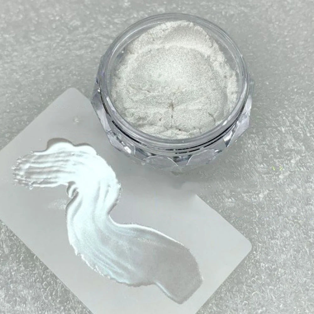 A small glass jar of silver glitter powder next to a white card showing a hand-painted white design.