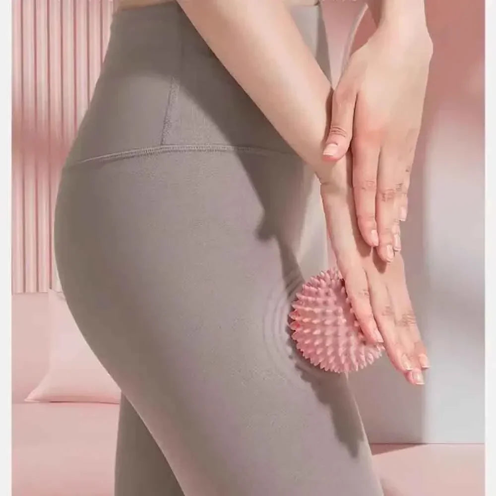 Close-up of a hand holding a pink deep tissue massage ball against a gray yoga pant leg.