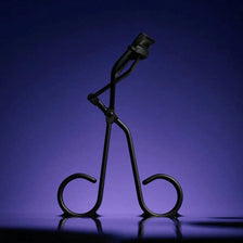 A black solid color metal eyelash curler is shown against a dark purple background.