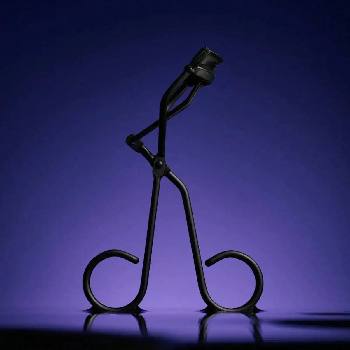 A black solid color metal eyelash curler is shown against a dark purple background.