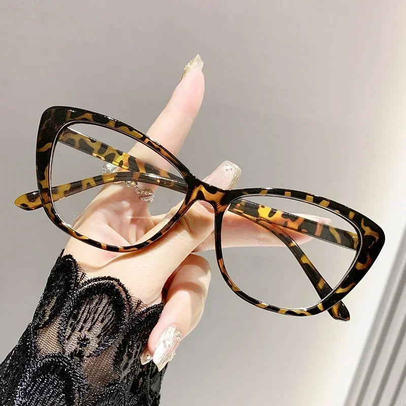A hand holds a pair of tortoiseshell cat eye reading glasses against a light background.