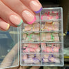 Une main tient une boîte en plastique transparent remplie de faux ongles acryliques colorés, dans différentes nuances de rose