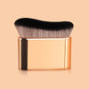 A rectangular gold-toned makeup brush with a black handle and angled wavy bristles resting on a beige background.