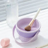 A purple silicone makeup brush cleaner pot with a wooden brush and a glass bottle are shown on a white surface.