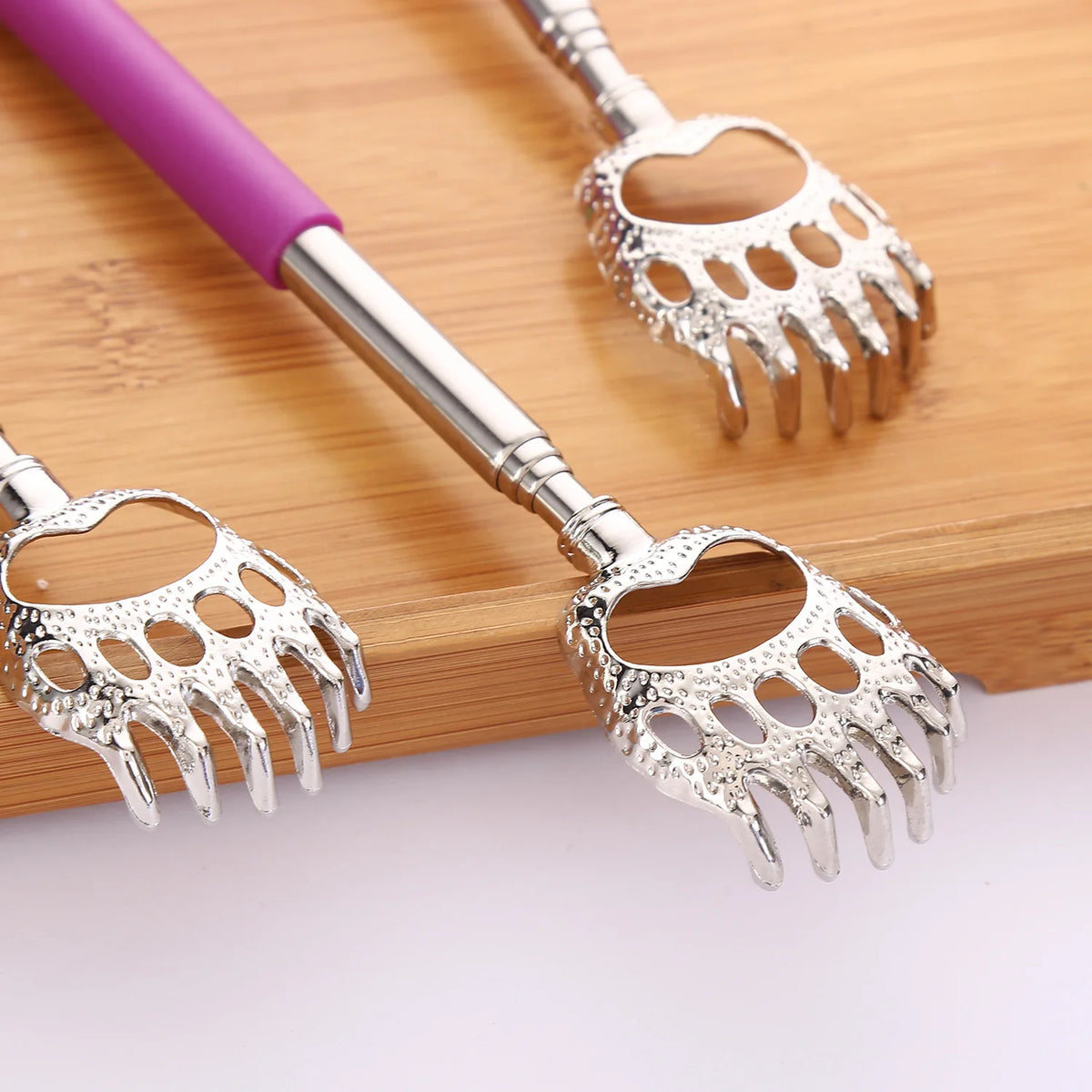 A stainless steel telescopic back scratcher with a purple handle and silver serrated fingers resting on a wooden surface.