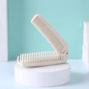 A white portable folding hairbrush with anti-static bristles resting on a white stand against a light blue background.