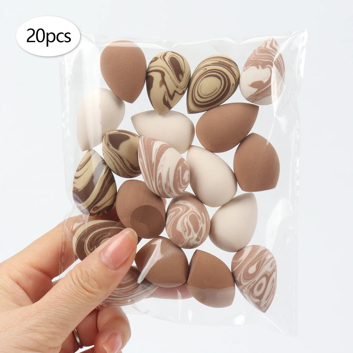 A hand holds a clear plastic bag containing 20 reusable expandable makeup sponges with marbled brown and beige patterns.