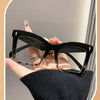 A hand holds a pair of black square frame reading glasses with clear lenses against a light background.