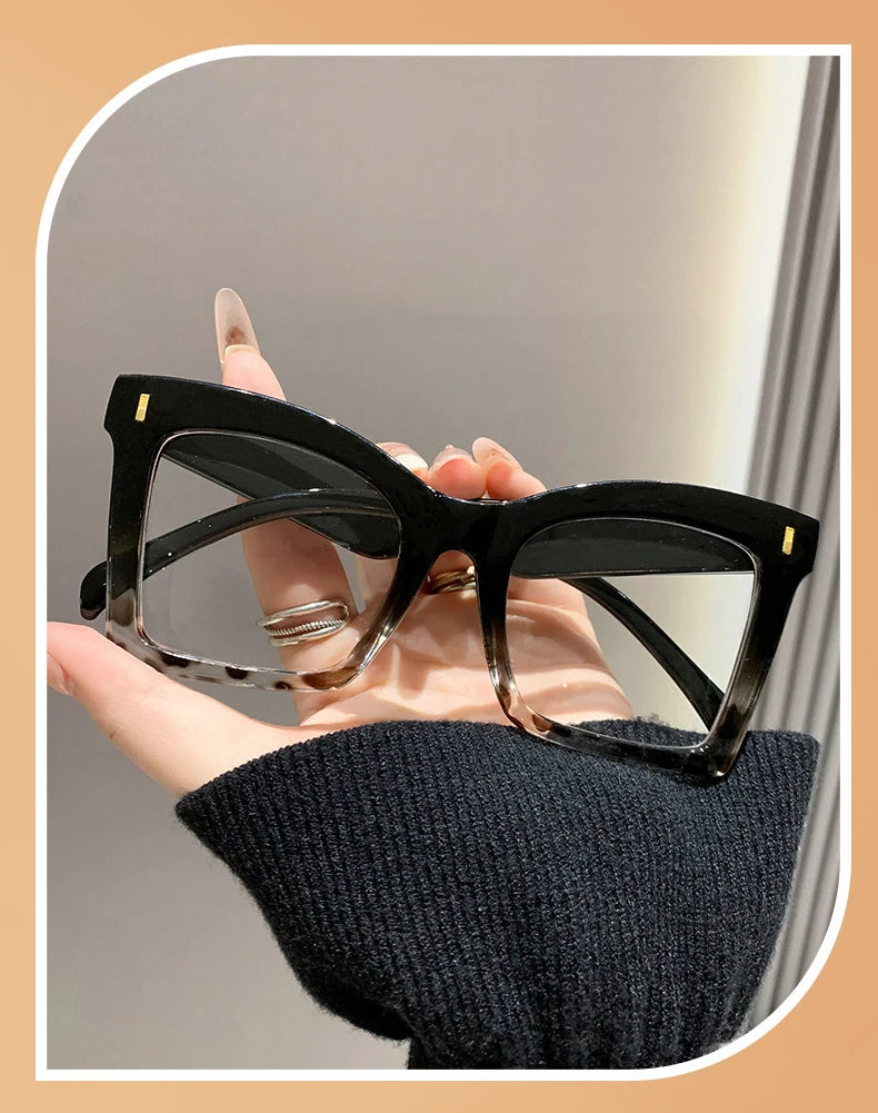A hand holds a pair of black square frame reading glasses with clear lenses against a light background.