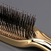 A close-up of a gold-colored brush with numerous thin, dark bristles used for scalp massage or hair styling.