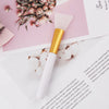 A gold and white silicone facial mask applicator brush resting on a pink paper background.