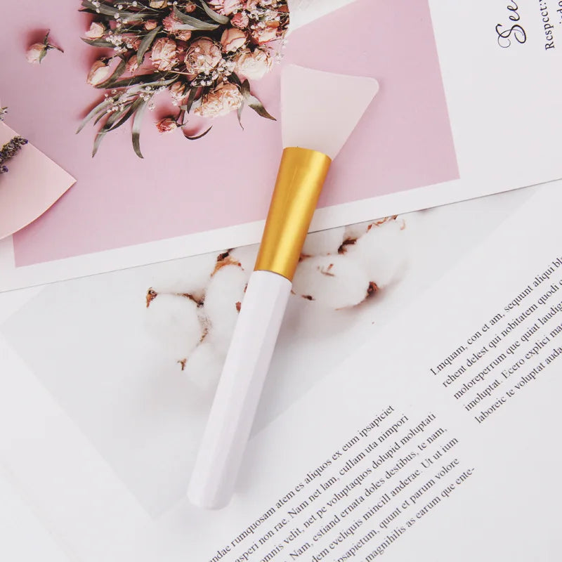 A gold and white silicone facial mask applicator brush resting on a pink paper background.