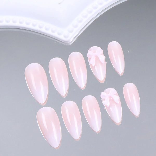 A set of pink almond press-on nails featuring a delicate bow design displayed on a gray surface.