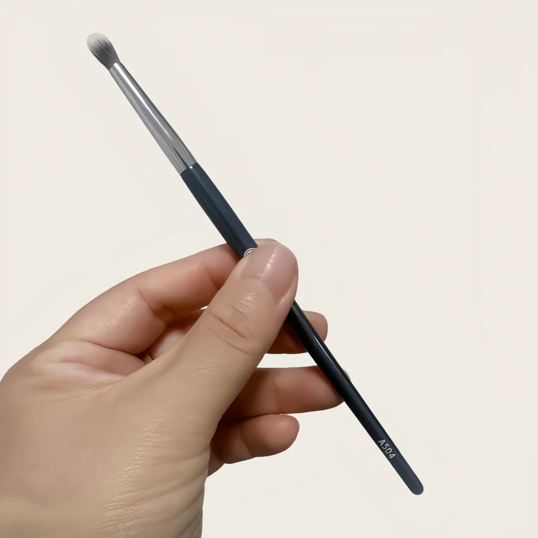 A hand holds a black precision concealer brush with a silver ferrule and a tapered tip.
