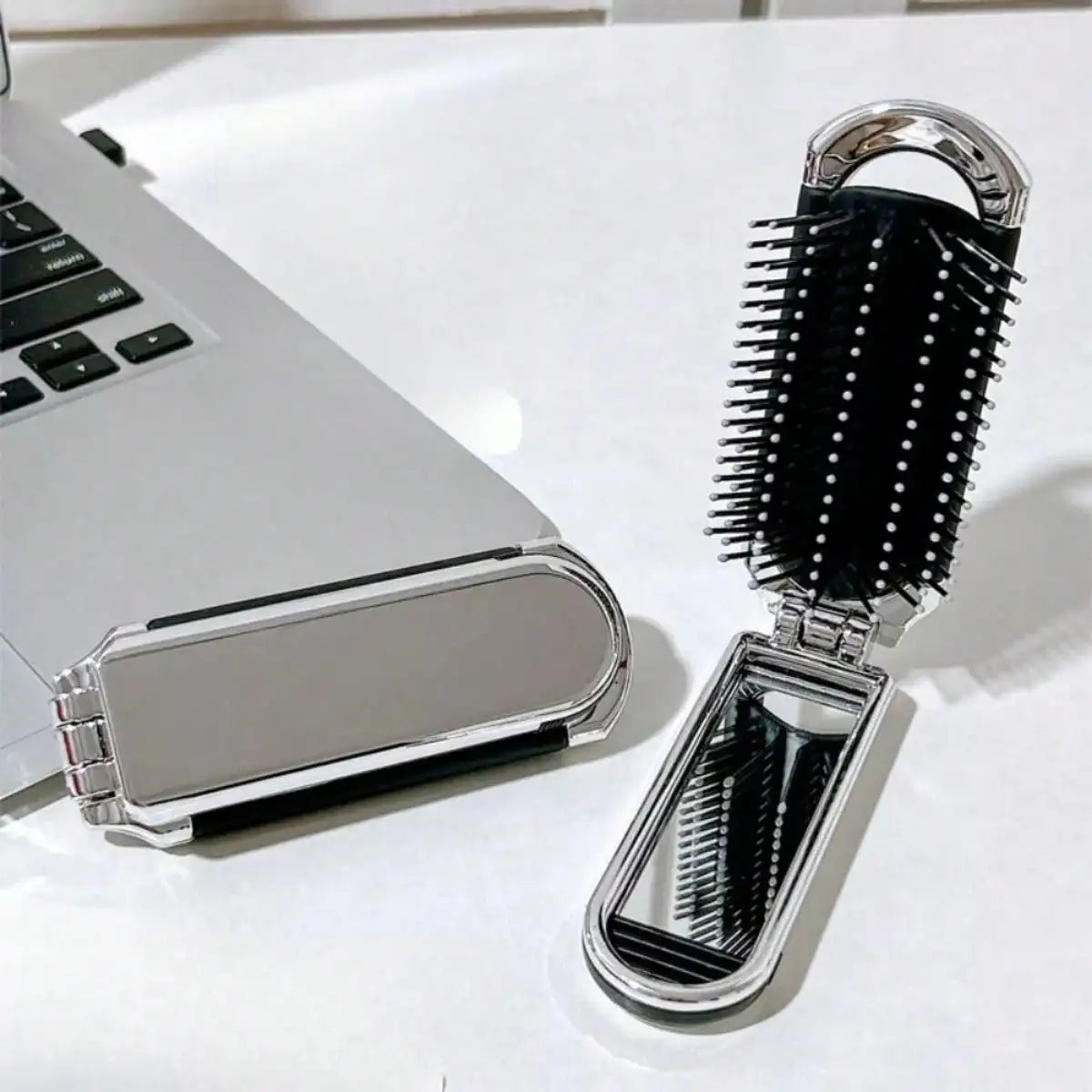 A silver portable compact comb with a black silicone bag and mirror is shown next to a laptop.